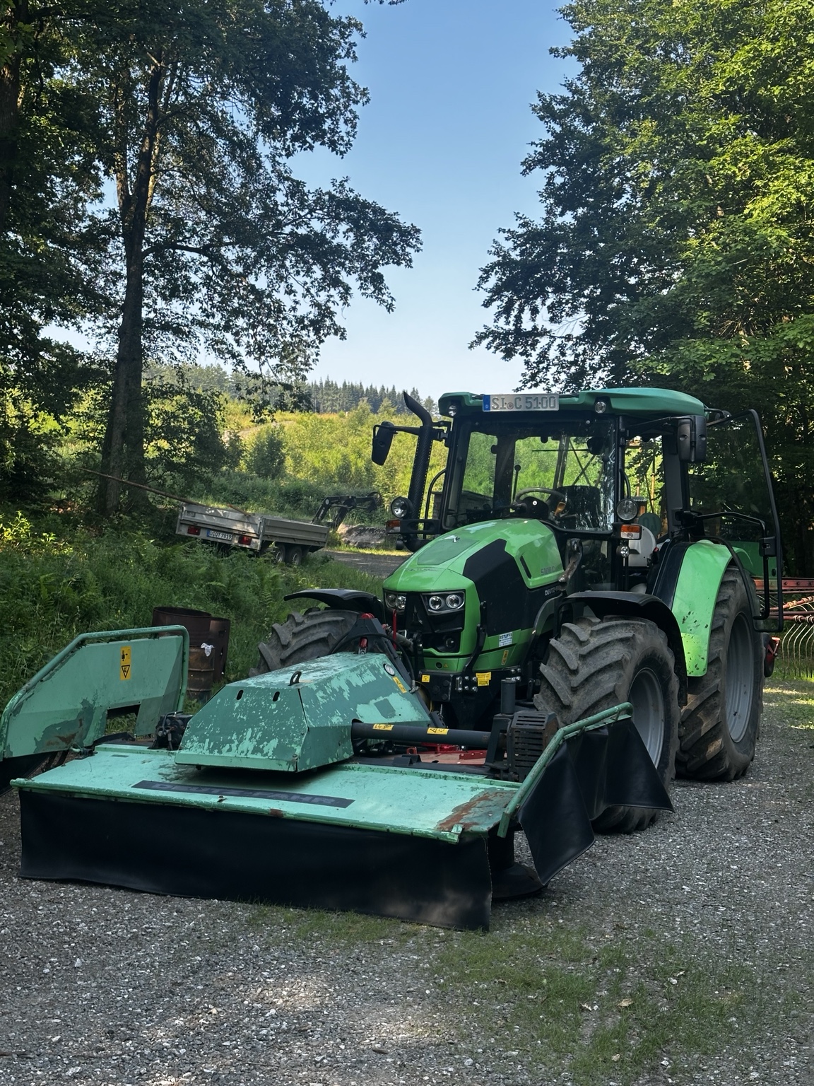 Grüner Traktor mit Frontmähwerk und Schwader am Waldrand
